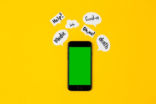 A smartphone with a green screen lies on a bright yellow background, surrounded by speech bubbles containing words like 'Help!', 'Covid-19', 'Oh no!', 'death', and 'Medic'. The arrangement suggests an alarming or urgent context.