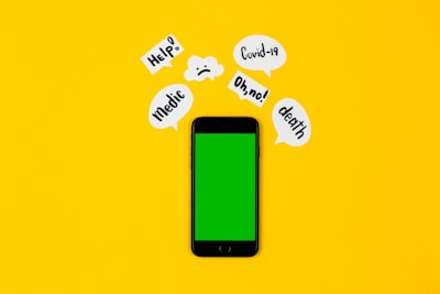 A smartphone with a green screen lies on a bright yellow background, surrounded by speech bubbles containing words like 'Help!', 'Covid-19', 'Oh no!', 'death', and 'Medic'. The arrangement suggests an alarming or urgent context.