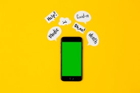 A smartphone with a green screen lies on a bright yellow background, surrounded by speech bubbles containing words like 'Help!', 'Covid-19', 'Oh no!', 'death', and 'Medic'. The arrangement suggests an alarming or urgent context.
