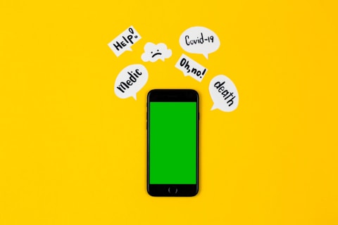 A smartphone with a green screen lies on a bright yellow background, surrounded by speech bubbles containing words like 'Help!', 'Covid-19', 'Oh no!', 'death', and 'Medic'. The arrangement suggests an alarming or urgent context.