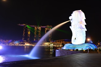 A night view of an iconic waterfront area featuring a large Merlion statue surrounded by bright lights, with a backdrop of modern skyscrapers, including a uniquely designed building with a sky park on top. Green laser lights are visible in the sky, and the water reflects the illuminated surroundings.