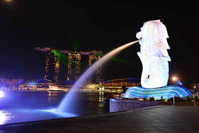 A night view of an iconic waterfront area featuring a large Merlion statue surrounded by bright lights, with a backdrop of modern skyscrapers, including a uniquely designed building with a sky park on top. Green laser lights are visible in the sky, and the water reflects the illuminated surroundings.