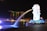 A night view of an iconic waterfront area featuring a large Merlion statue surrounded by bright lights, with a backdrop of modern skyscrapers, including a uniquely designed building with a sky park on top. Green laser lights are visible in the sky, and the water reflects the illuminated surroundings.