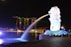 A night view of an iconic waterfront area featuring a large Merlion statue surrounded by bright lights, with a backdrop of modern skyscrapers, including a uniquely designed building with a sky park on top. Green laser lights are visible in the sky, and the water reflects the illuminated surroundings.