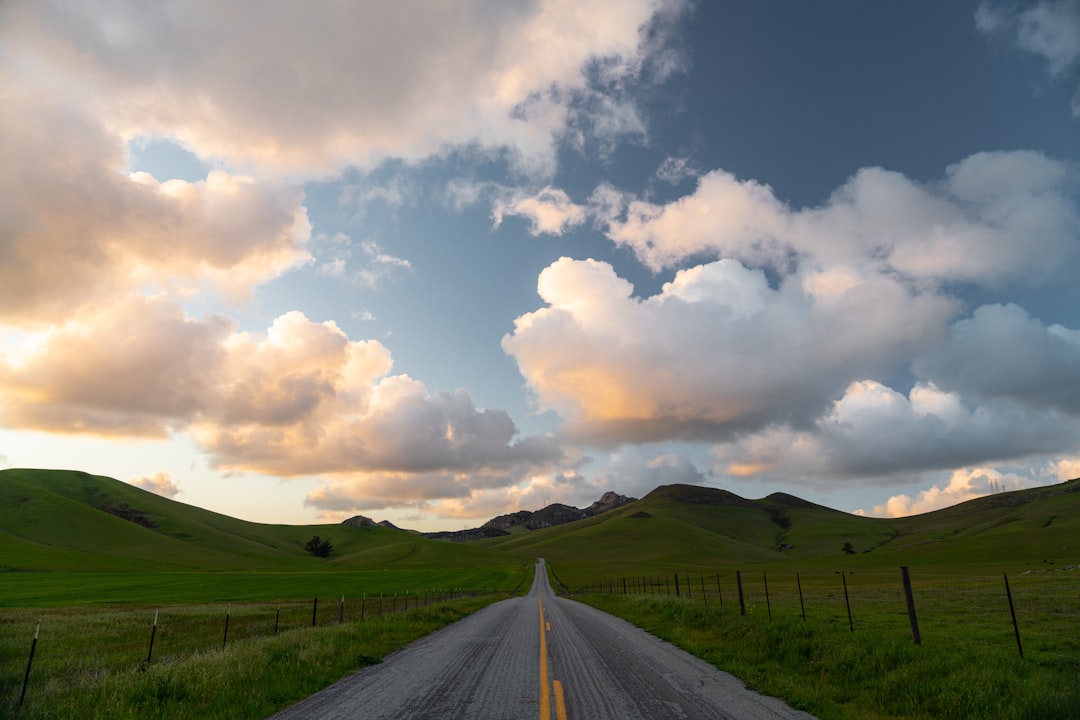 gray asphalt road between green grass field under white clouds and blue sky during daytime