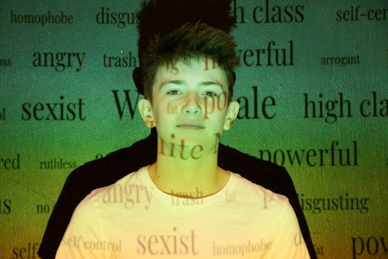 A young person stands against a background filled with various words like 'angry,' 'powerful,' and 'sexist' projected onto them. The lighting casts a green and yellowish hue across the scene, creating a contrast with the person's white shirt.
