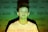 A young person stands against a background filled with various words like 'angry,' 'powerful,' and 'sexist' projected onto them. The lighting casts a green and yellowish hue across the scene, creating a contrast with the person's white shirt.