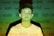 A young person stands against a background filled with various words like 'angry,' 'powerful,' and 'sexist' projected onto them. The lighting casts a green and yellowish hue across the scene, creating a contrast with the person's white shirt.