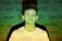 A young person stands against a background filled with various words like 'angry,' 'powerful,' and 'sexist' projected onto them. The lighting casts a green and yellowish hue across the scene, creating a contrast with the person's white shirt.