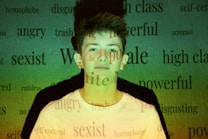 A young person stands against a background filled with various words like 'angry,' 'powerful,' and 'sexist' projected onto them. The lighting casts a green and yellowish hue across the scene, creating a contrast with the person's white shirt.