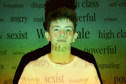 A young person stands against a background filled with various words like 'angry,' 'powerful,' and 'sexist' projected onto them. The lighting casts a green and yellowish hue across the scene, creating a contrast with the person's white shirt.
