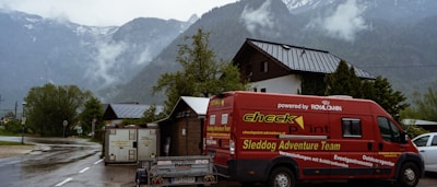 A vibrant red van with the words 'Sleddog Adventure Team' is parked on a scenic road set against a backdrop of misty mountains. The area is damp, indicating recent rain, and surrounded by lush greenery and rustic buildings.