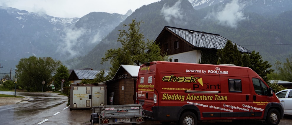 A vibrant red van with the words 'Sleddog Adventure Team' is parked on a scenic road set against a backdrop of misty mountains. The area is damp, indicating recent rain, and surrounded by lush greenery and rustic buildings.