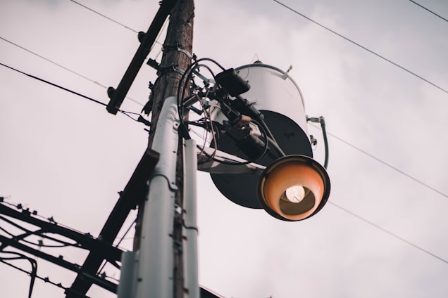 A utility pole with an attached transformer and an industrial-style streetlight. The sky is overcast, providing a muted backdrop. Electrical wires extend outward, creating a network across the scene.