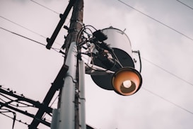 A utility pole with an attached transformer and an industrial-style streetlight. The sky is overcast, providing a muted backdrop. Electrical wires extend outward, creating a network across the scene.