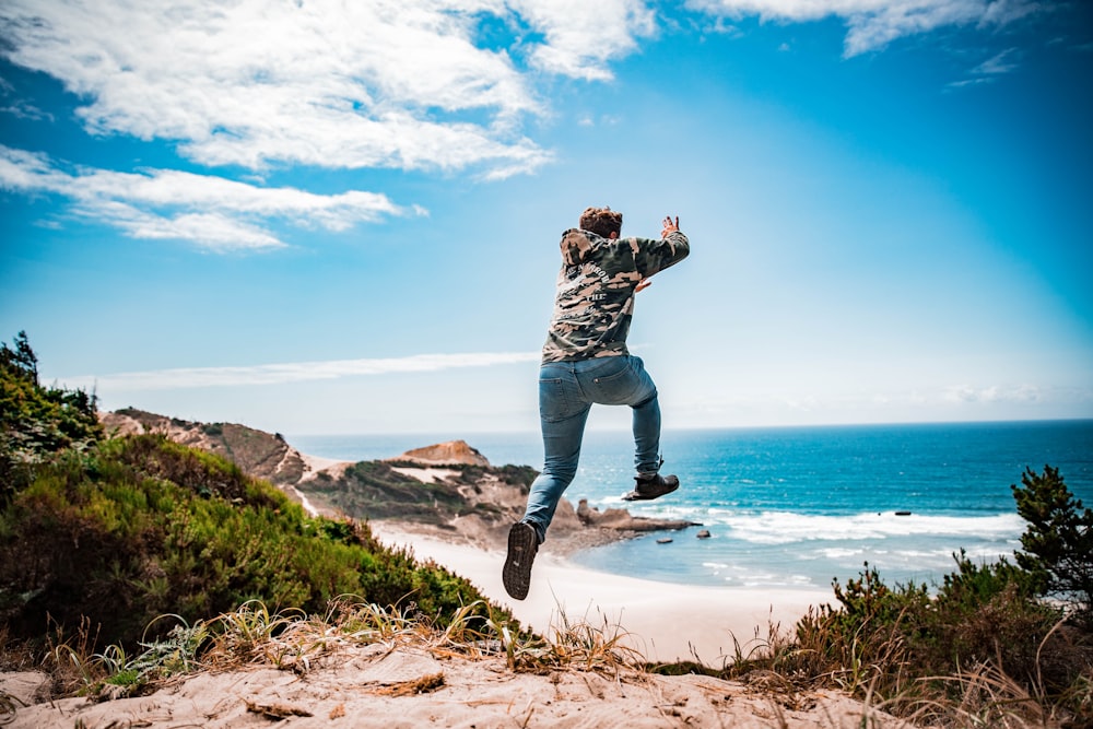 man in black and white camouflage jacket and blue denim jeans jumping on brown rock formation