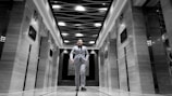 A man dressed in a suit walks confidently down the center of an elegant corridor with modern design elements. The ceiling features an artistic wave-like pattern with light fixtures. The walls on either side have a glossy finish with embedded digital screens, and there are elevator doors.
