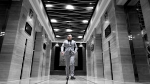 A man dressed in a suit walks confidently down the center of an elegant corridor with modern design elements. The ceiling features an artistic wave-like pattern with light fixtures. The walls on either side have a glossy finish with embedded digital screens, and there are elevator doors.