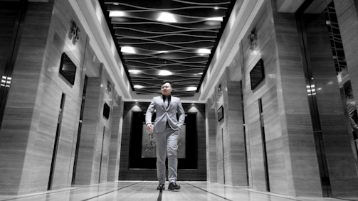 A man dressed in a suit walks confidently down the center of an elegant corridor with modern design elements. The ceiling features an artistic wave-like pattern with light fixtures. The walls on either side have a glossy finish with embedded digital screens, and there are elevator doors.
