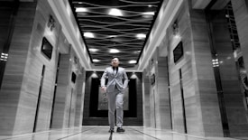 A man dressed in a suit walks confidently down the center of an elegant corridor with modern design elements. The ceiling features an artistic wave-like pattern with light fixtures. The walls on either side have a glossy finish with embedded digital screens, and there are elevator doors.