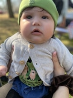 A baby wearing a green hat and a light-colored cardigan is gazing upwards. The cardigan features wooden buttons and underneath, there's a shirt with forest-themed designs and animals. The baby is being held by someone with polished nails wearing a dark hair scrunchie. The background suggests an outdoor setting with blurred trees and grass.