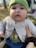 A baby wearing a green hat and a light-colored cardigan is gazing upwards. The cardigan features wooden buttons and underneath, there's a shirt with forest-themed designs and animals. The baby is being held by someone with polished nails wearing a dark hair scrunchie. The background suggests an outdoor setting with blurred trees and grass.