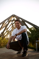 A person squatting on a wooden platform with a bright smile, wearing a white t-shirt and dark jeans. A distinctive triangular structure is visible in the background, and lush greenery surrounds the area. The lighting suggests a warm, sunny day.