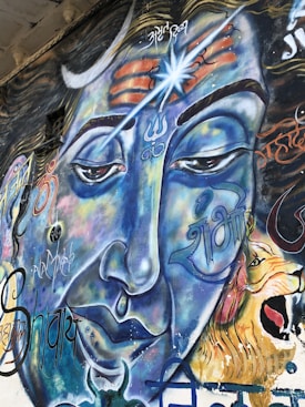 A vibrant mural of a deity displays detailed facial features with closed eyes and a calm expression. The face is dominated by blue hues, with intricate symbols and patterns around the forehead, including a trident and a third eye motif. Script and abstract designs are incorporated around the visage. A lion, depicted with an open mouth, is positioned on the right side of the image.