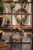 A rustic interior setting features an assortment of hanging wreaths and plants against an old window frame backdrop. The wreaths are decorated with a variety of succulents, moss, and dried flowers, adding a natural and earthy touch. A woven basket is visible on the side, enhancing the organic theme.