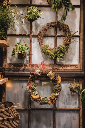 A rustic interior setting features an assortment of hanging wreaths and plants against an old window frame backdrop. The wreaths are decorated with a variety of succulents, moss, and dried flowers, adding a natural and earthy touch. A woven basket is visible on the side, enhancing the organic theme.