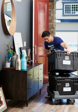 A person wearing a blue t-shirt is inside a house, handling two large black containers labeled 'Hive Boxx' on a wheeled dolly. The person is standing near a dark wooden cabinet adorned with decorative items, including framed pictures and a plant. In the background, a partially open red door and exterior window are visible.