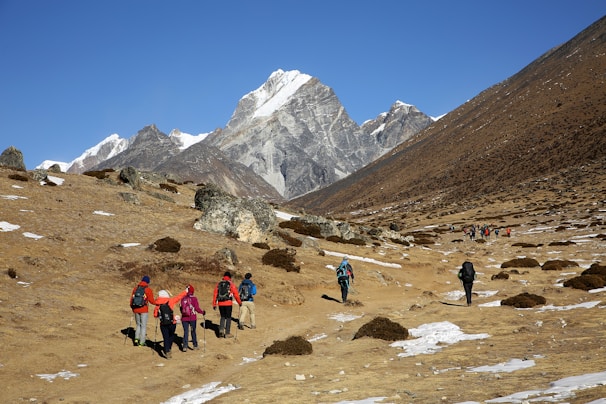 A group of hikers is walking along a rocky trail in a mountainous region. Snow-capped peaks rise majestically in the distance under a clear blue sky. The landscape is barren with patches of snow and small shrubs scattered across the rocky terrain.