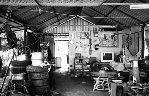 A cluttered garage-like space with a corrugated metal roof. Stacks of old tires and metal rims are positioned on the left. A small television is placed on a table at the rear against a wall covered with various items, including a painting or poster of horses. The room is filled with various mechanical parts and tools scattered around.