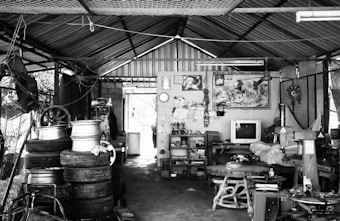 A cluttered garage-like space with a corrugated metal roof. Stacks of old tires and metal rims are positioned on the left. A small television is placed on a table at the rear against a wall covered with various items, including a painting or poster of horses. The room is filled with various mechanical parts and tools scattered around.