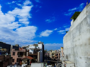 A vibrant cityscape with densely packed buildings showcasing a variety of architectural styles and colors under a bright blue sky with scattered white clouds. Satellite dishes and rooftop structures are visible, adding to the urban feel.