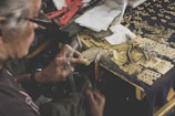 An elderly person is working on a detailed craft project at a cluttered workspace. The desk is covered in metal pieces, small tools, and various notes. The person is holding a tool in one hand and appears to be assembling or engraving a small metallic component.