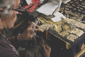 An elderly person is working on a detailed craft project at a cluttered workspace. The desk is covered in metal pieces, small tools, and various notes. The person is holding a tool in one hand and appears to be assembling or engraving a small metallic component.