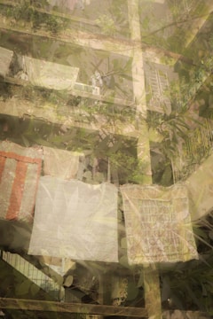 A multi-story residential building with lush greenery and plants on the balconies. Laundry is hung out to dry, including large sheets and possibly blankets. The structure appears to be aged with visible concrete, and the atmosphere is peaceful with a sense of everyday life.