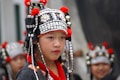 A child wearing a traditional outfit adorned with intricate beads and decorations stands prominently. The headpiece features black fabric with white beads, silver ornaments, and red accents. In the background, more children are wearing similar attire, slightly out of focus.