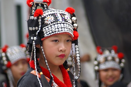 A child wearing a traditional outfit adorned with intricate beads and decorations stands prominently. The headpiece features black fabric with white beads, silver ornaments, and red accents. In the background, more children are wearing similar attire, slightly out of focus.