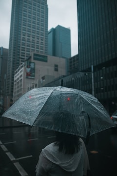 A person stands under a transparent umbrella in the rain, surrounded by tall buildings in an urban setting. Raindrops are visible on the umbrella, and the streets appear wet and reflective from the rain.