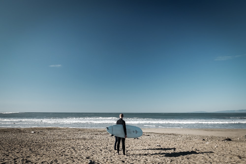 man in white shirt carrying white surfboard walking on beach during daytime