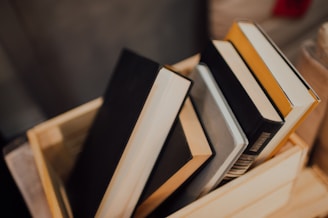 A wooden box contains several hardcover books with different colored covers. The books are stacked at an angle inside the box, with covers in black, gray, and yellow. The background is blurred, focusing attention on the books.