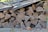 A neatly stacked pile of cut firewood is sheltered under a corrugated metal roof. The wood logs are arranged in a compact manner with visible growth rings and bark, showing different shades of brown and grey. Some dried leaves and moss can be seen around the base, adding to the rustic outdoor setting.