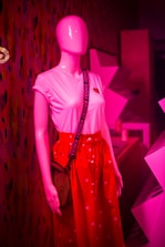 A pink mannequin is dressed in a white t-shirt with a heart emblem and a bright red skirt adorned with small white patterns. The mannequin is also wearing a crossbody bag with a black strap that has the word 'BAGGU' printed repeatedly. The background consists of geometric shapes and patterns lit in pink lighting.