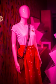 A pink mannequin is dressed in a white t-shirt with a heart emblem and a bright red skirt adorned with small white patterns. The mannequin is also wearing a crossbody bag with a black strap that has the word 'BAGGU' printed repeatedly. The background consists of geometric shapes and patterns lit in pink lighting.