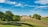 A picturesque landscape featuring a rustic stone farmhouse situated on a grassy hill. The house has a red-tiled roof and is surrounded by trees and open fields under a bright blue sky with scattered clouds.
