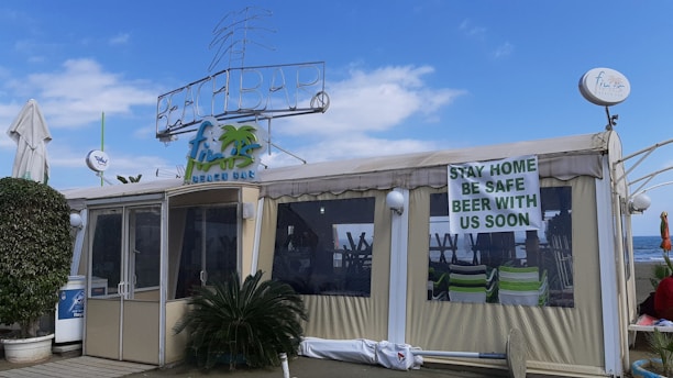A beach bar structure with a sign on top reading 'BEACH BAR' and another indicating the name with a palm tree illustration. The front has several glass windows and a door, with a large banner saying 'STAY HOME BE SAFE BEER WITH US SOON'. There are tropical plants and umbrellas around, and the sea is visible in the background under a partly cloudy sky.