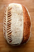 A beautifully crafted loaf of sourdough bread with intricate scoring patterns on the crust, lying on a wooden surface. The bread has a crackling texture with a blend of light and dark brown hues, indicating a well-baked exterior.