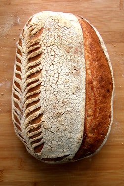 A beautifully crafted loaf of sourdough bread with intricate scoring patterns on the crust, lying on a wooden surface. The bread has a crackling texture with a blend of light and dark brown hues, indicating a well-baked exterior.